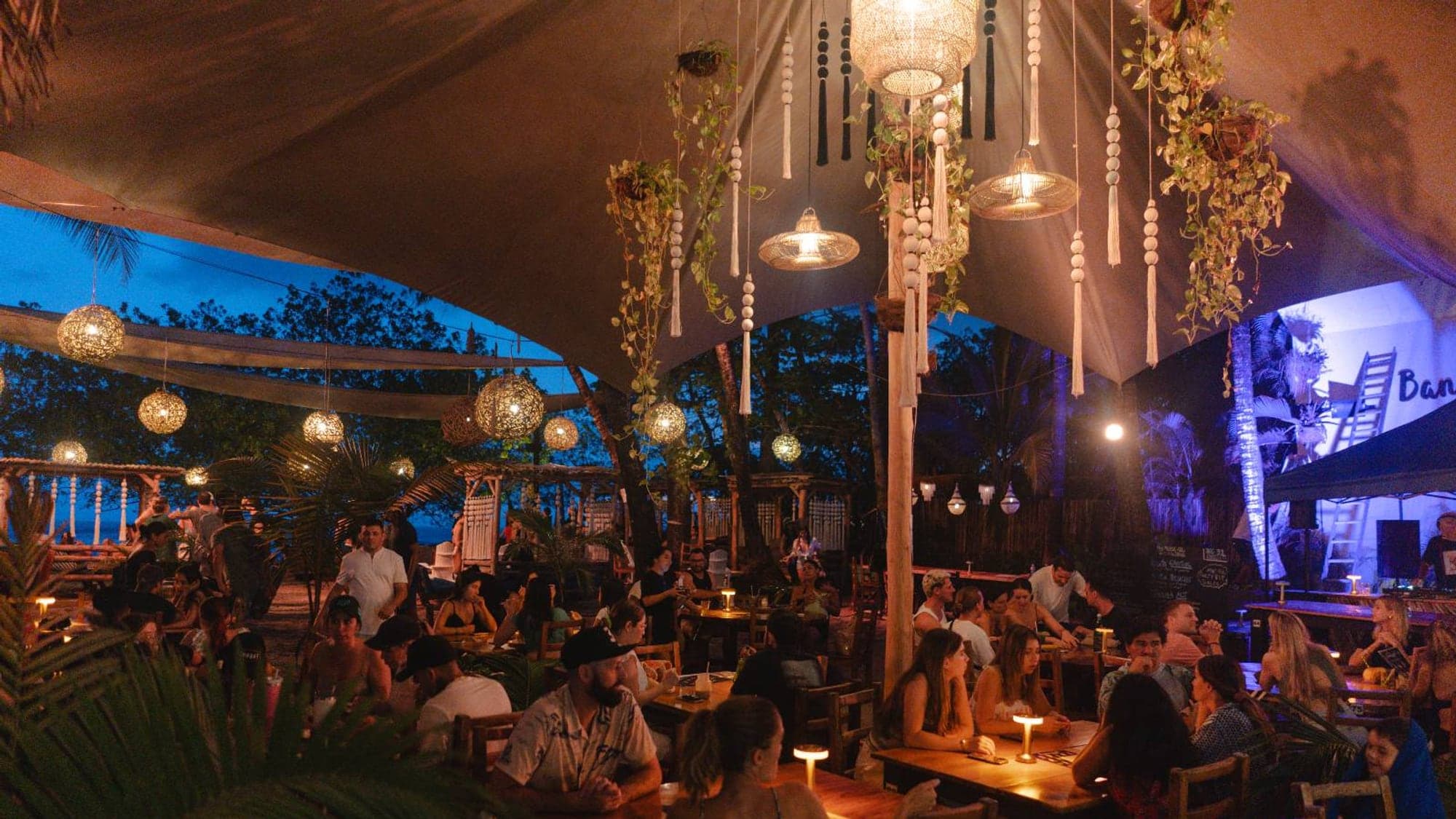 Open-air Santa Teresa beach bar at night with macrame lanterns, candle tables, and a packed crowd